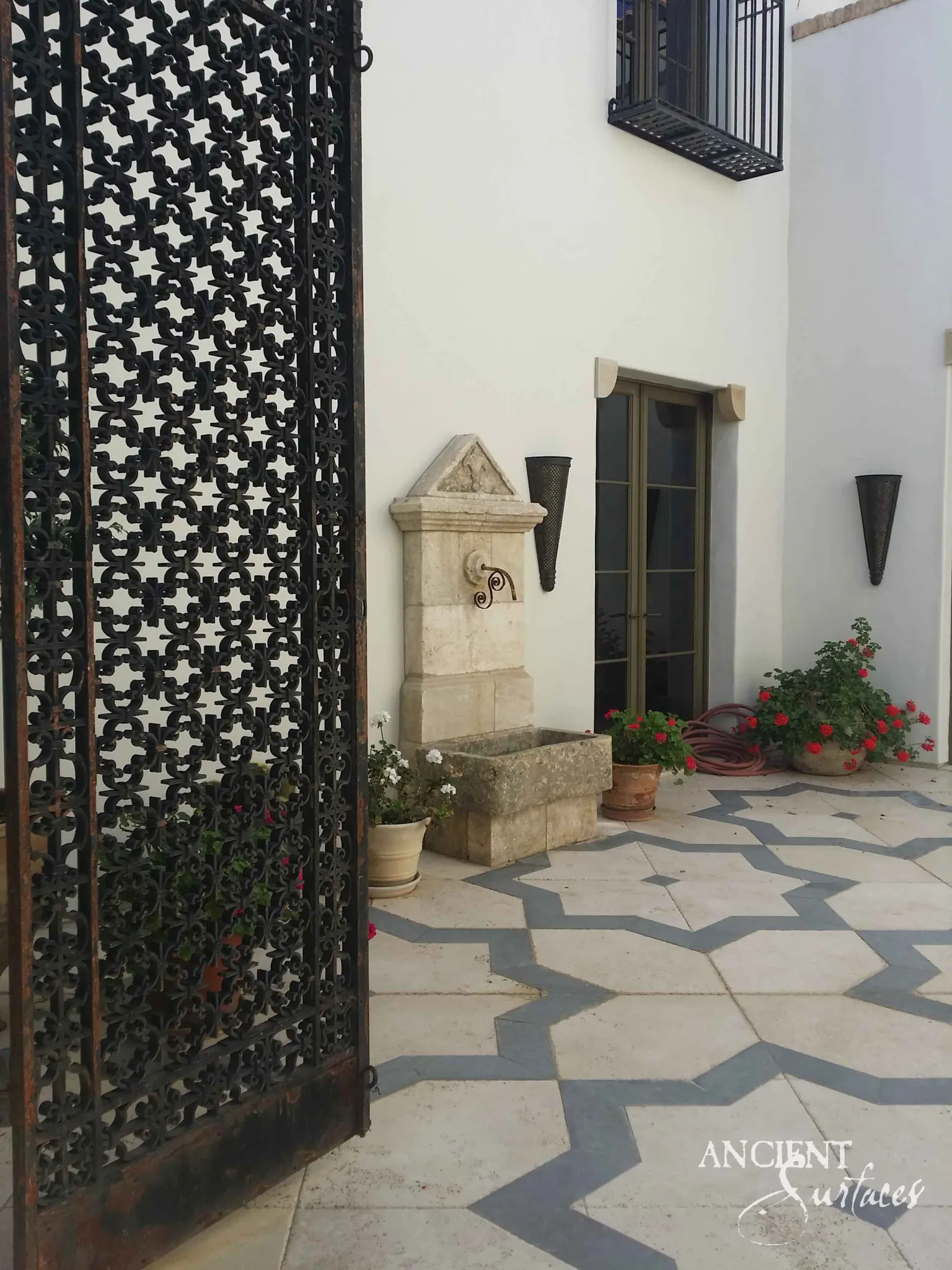 Antique limestone wall fountain set against a white courtyard wall beside an ornate iron gate